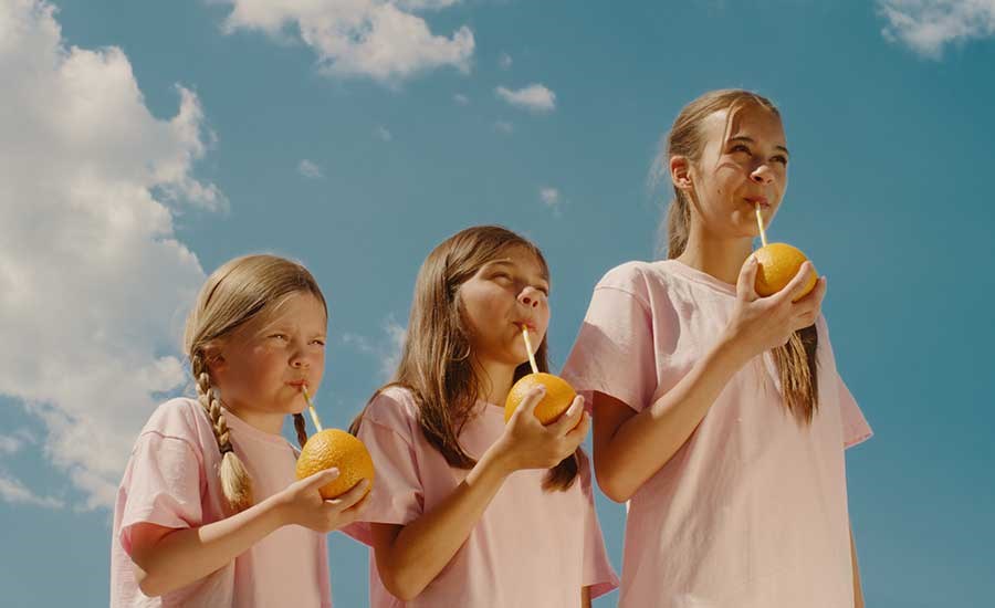 Tre barn tittar snett framåt och har på sig ljusrosa t-shirt och håller varsin apelsin med sugrör i